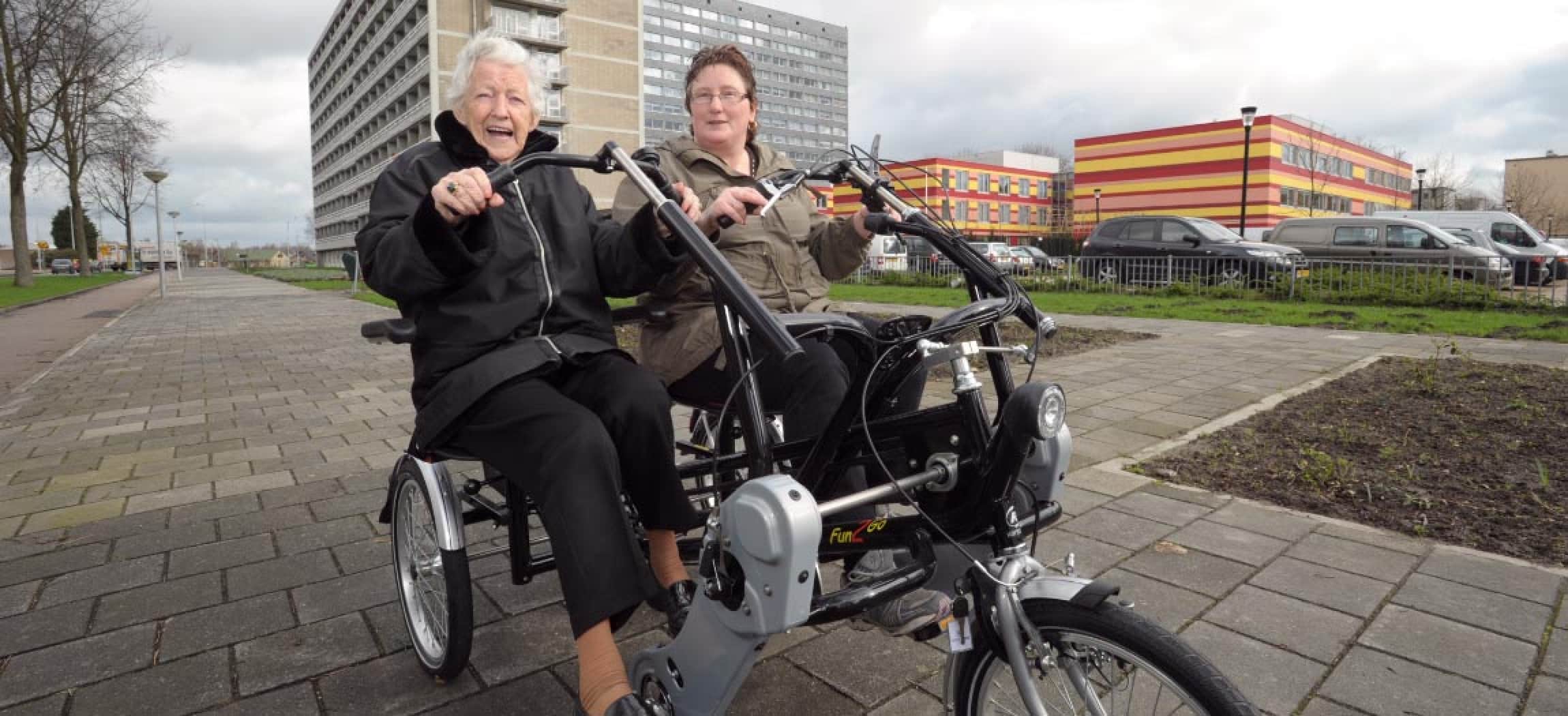Twee dames oma buiten op duofiets met gebouwen op achtergrond