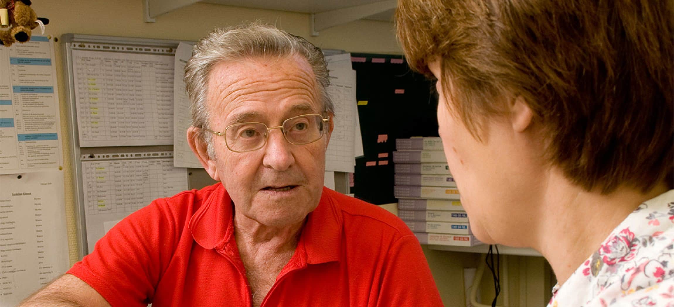 Man rood shirt bril kijkt en luistert naar logopedist met werkschema