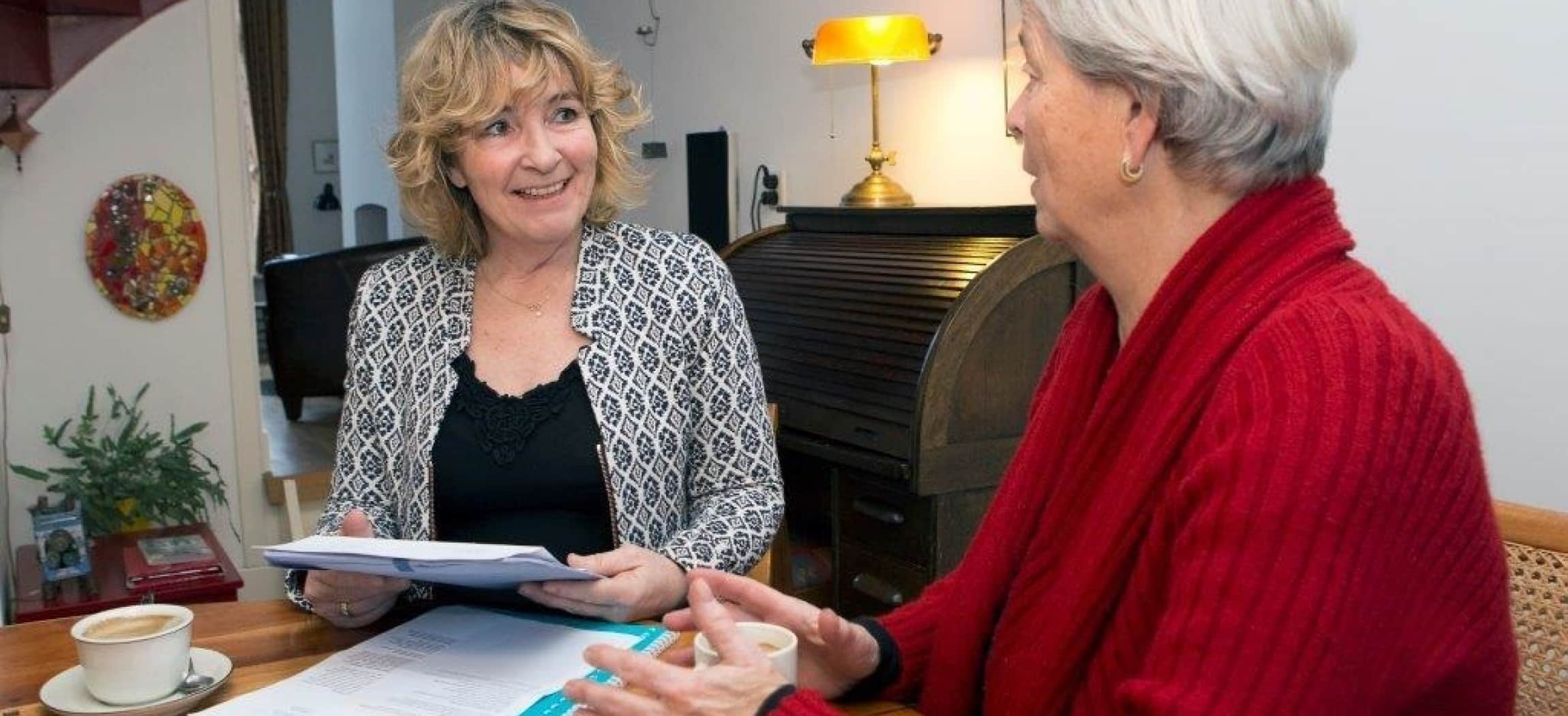 Twee vrouwen aan tafel met kopje koffie, de jongere vrouw houdt documenten vast en praat