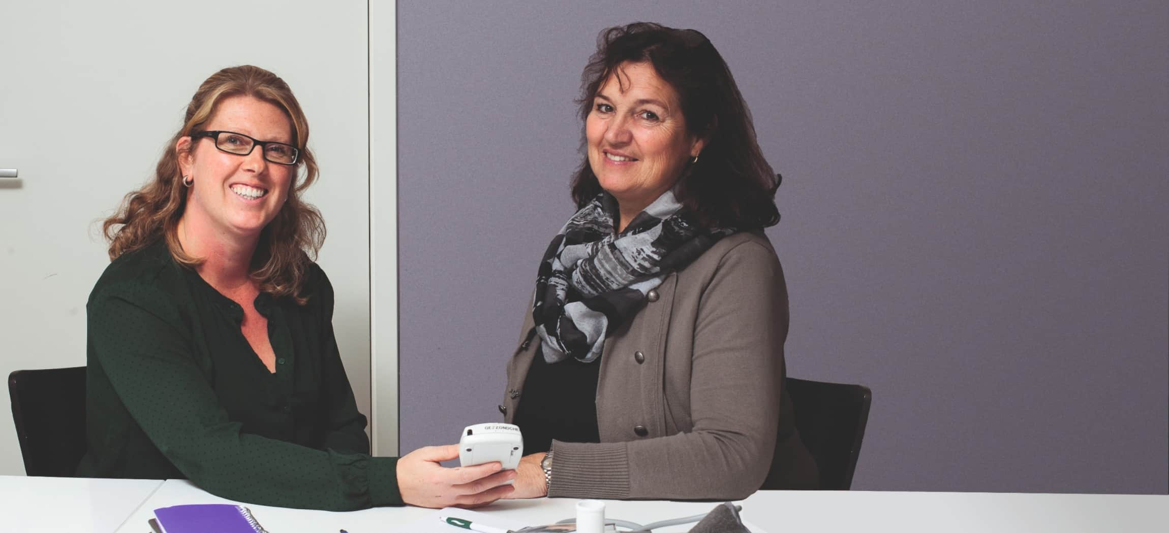 Twee vrouwen bekijken een boekje met poster Schijf van Vijf op achtergrond