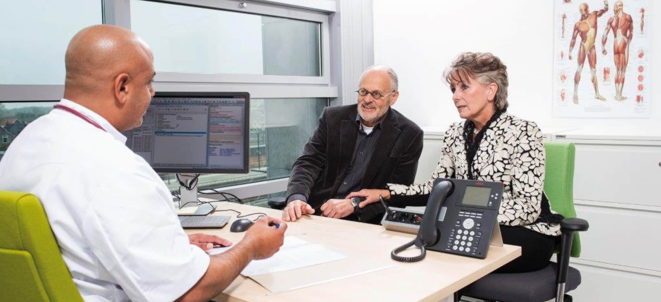 Man en vrouw praten met arts bij tafel met computer en telefoon, poster anatomie spieren lichaam