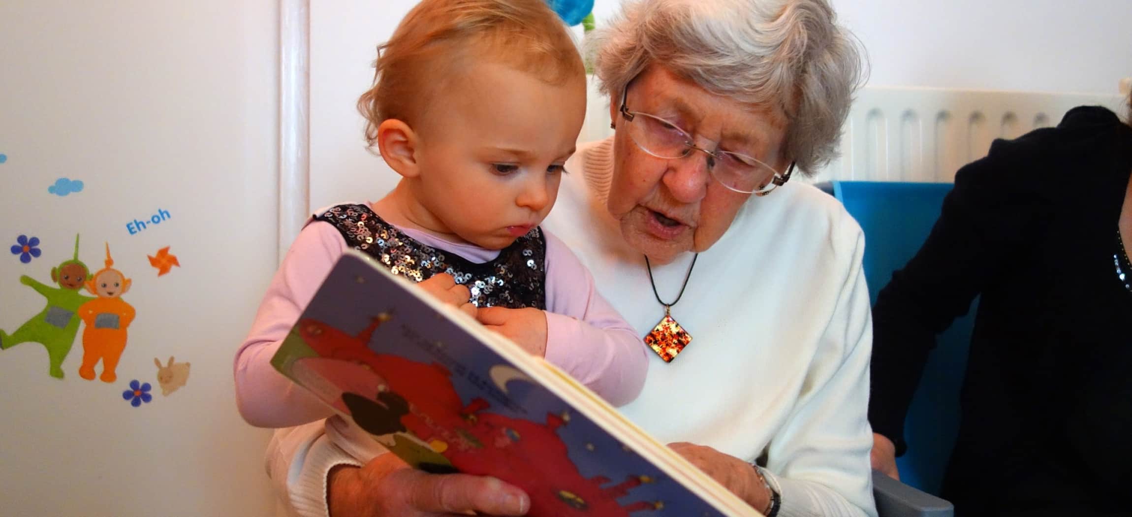 Meisje zit bij oma op schoot en oma leest voor uit een lees- en plaatjesboek
