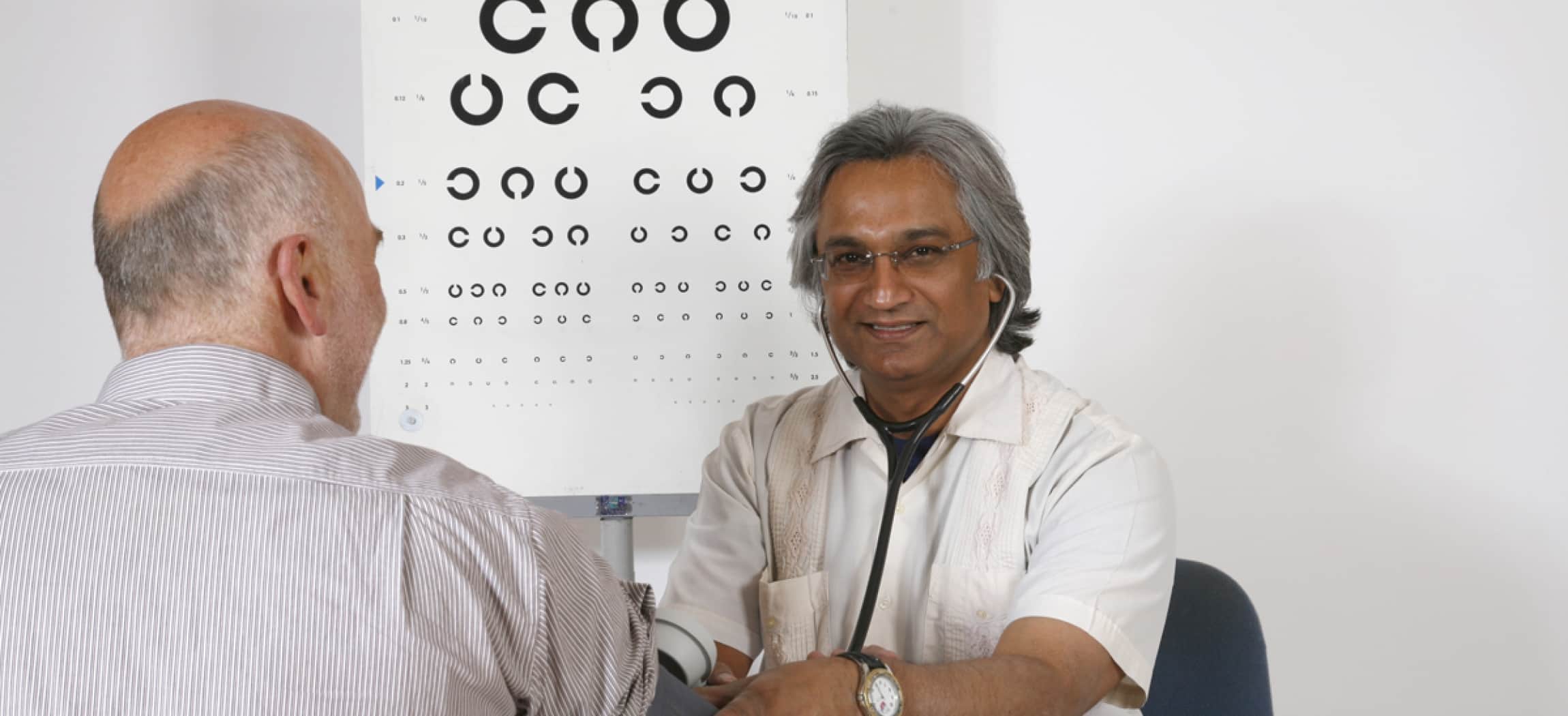 Arts doet een bloeddrukmeter om bij arm van man met poster oogtest