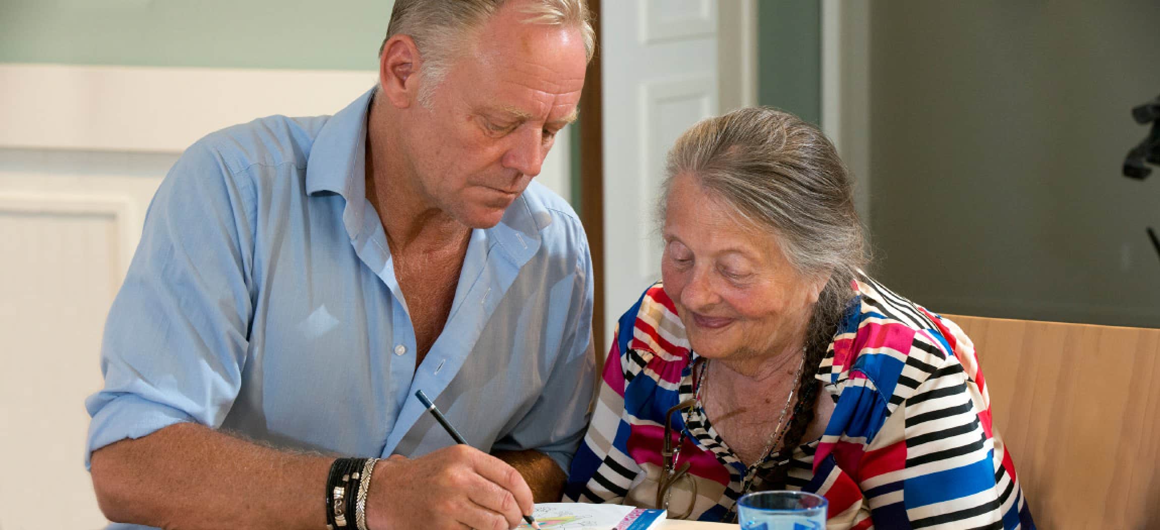 Man in lichtblauw overhemd kleurt een tekening voor bejaarde oma met dementie