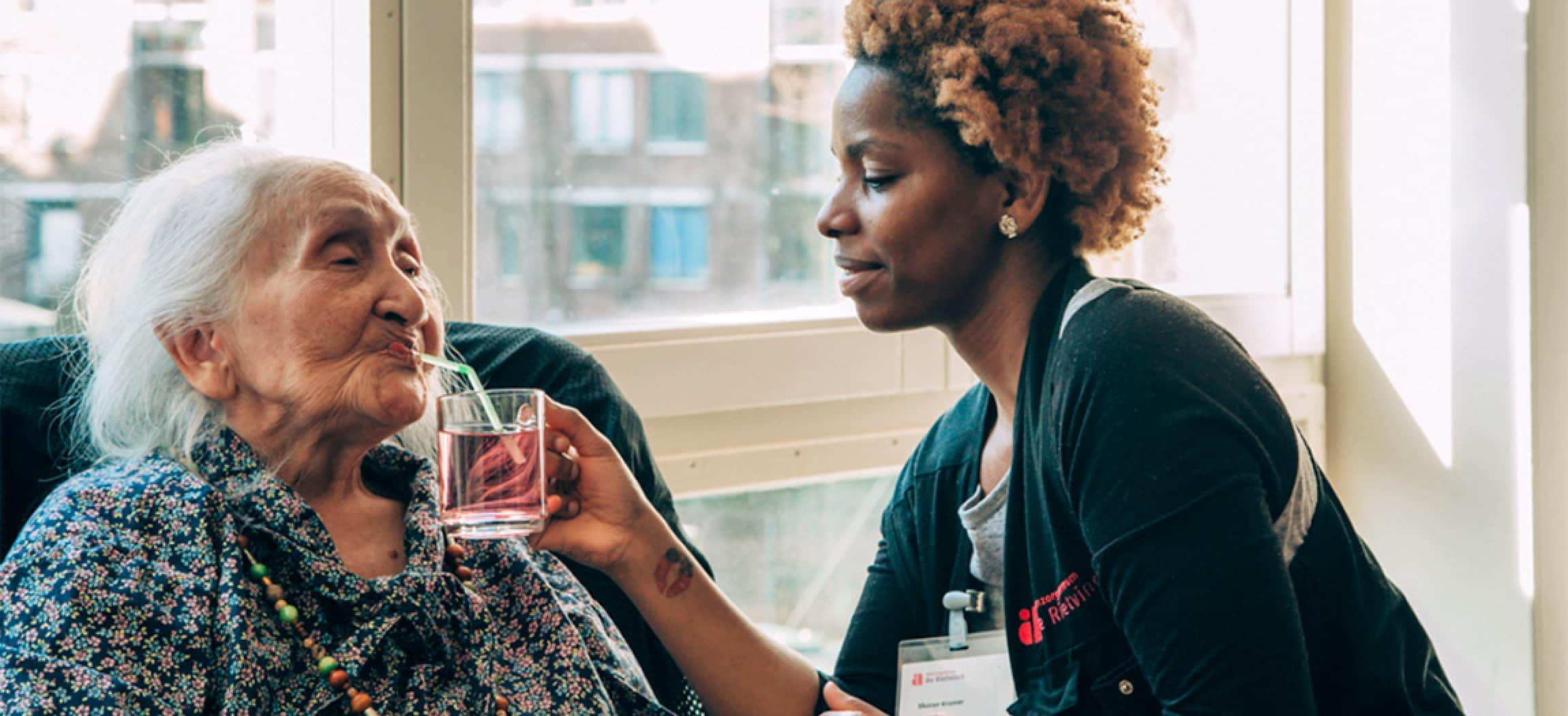 Verzorgende met krullend haar geeft oude vrouw drinken met rietje