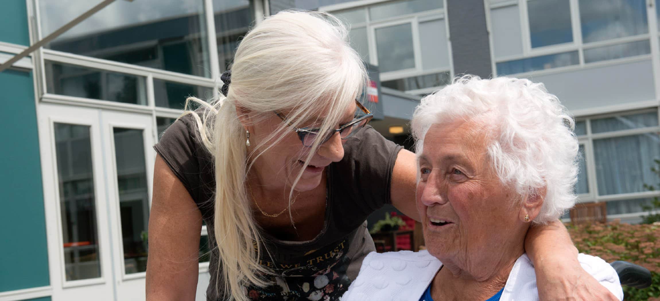 Vrouw met lang grijsblond haar omarmt bejaarde vrouw, gebouw op achtergrond