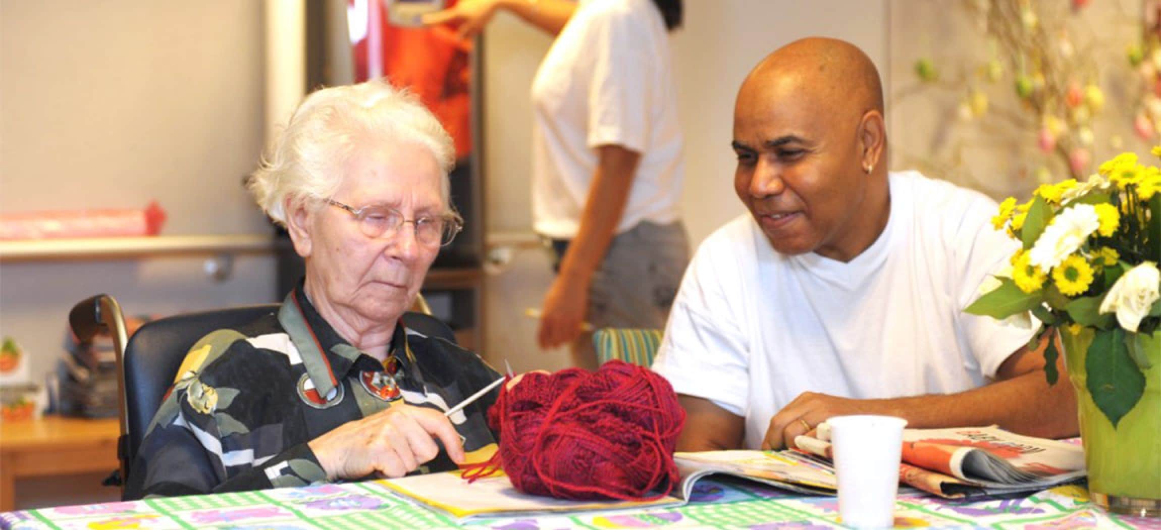 Mannelijke verzorgende kijkt naar oude vrouw met breiwerk en bol wol