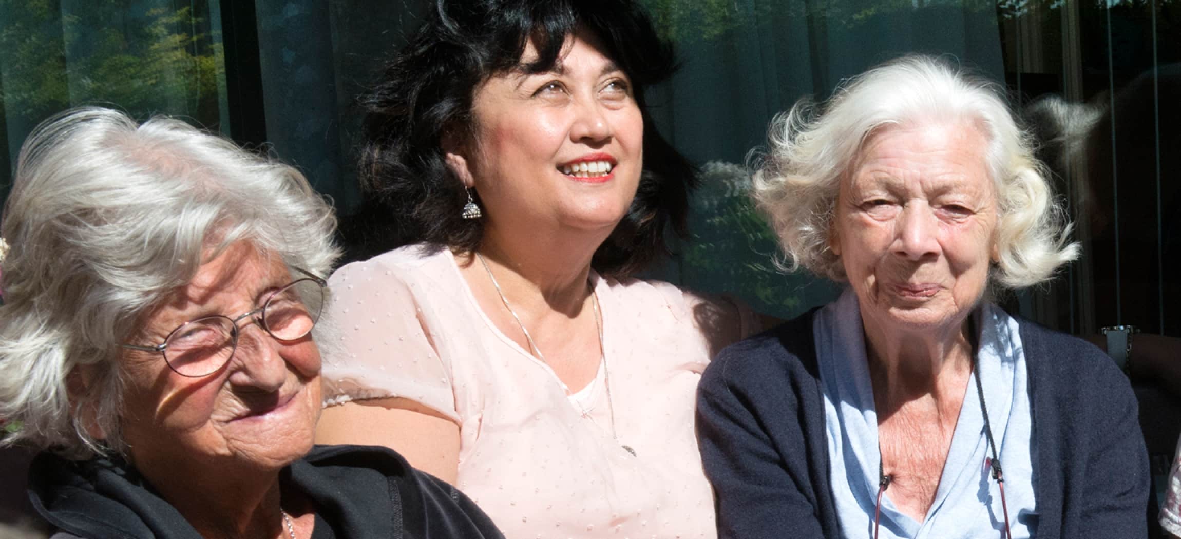 Vrouw met zwart haar en roze blouse zit tussen twee bejaarde dames