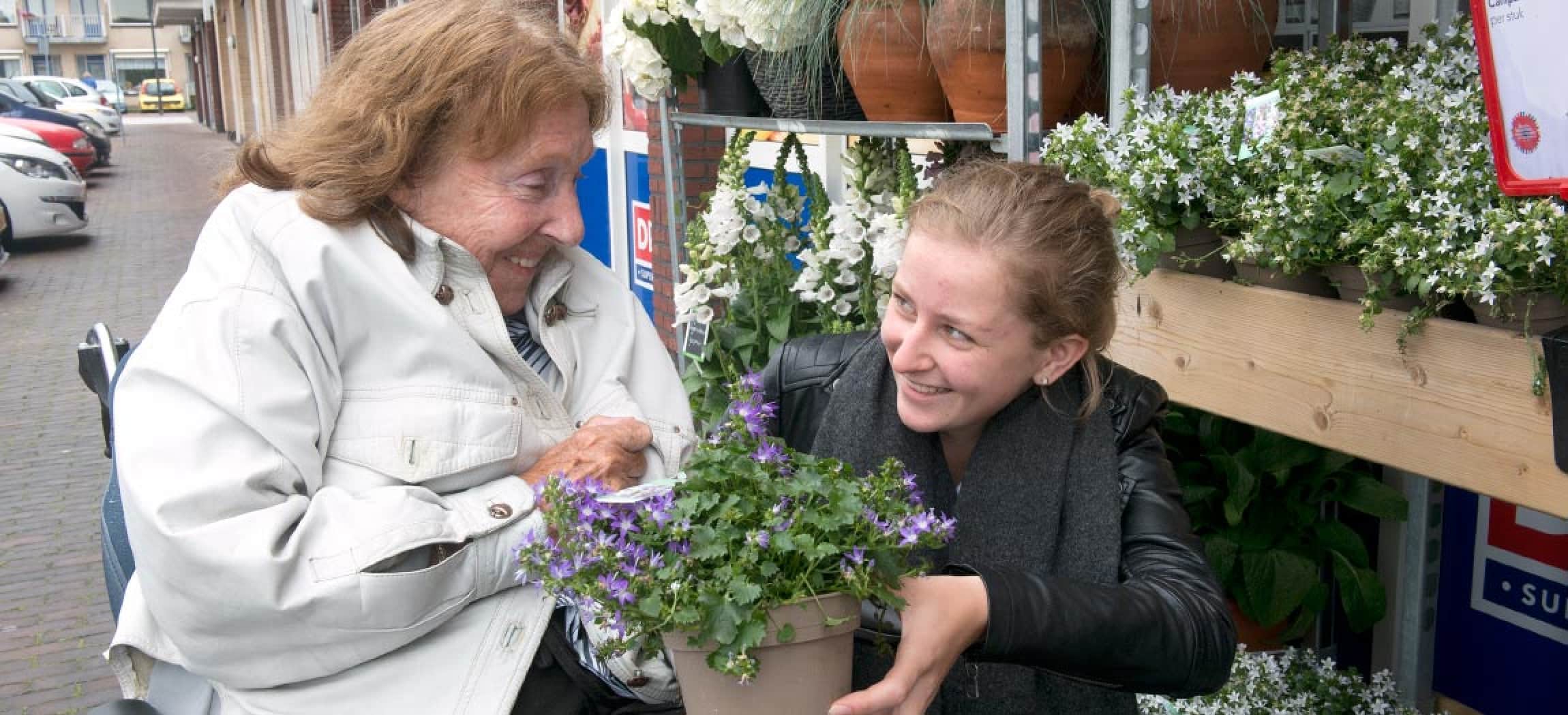 Stagiaire MBO laat bij winkels plantjes zien aan oudere vrouw in rolstoel