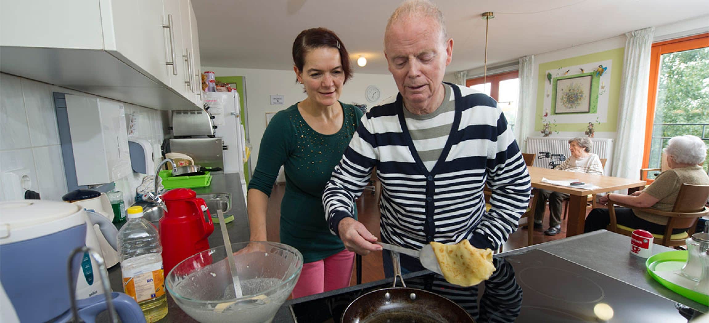 Oudere man bakt een pannenkoek met hulp van verzorgende