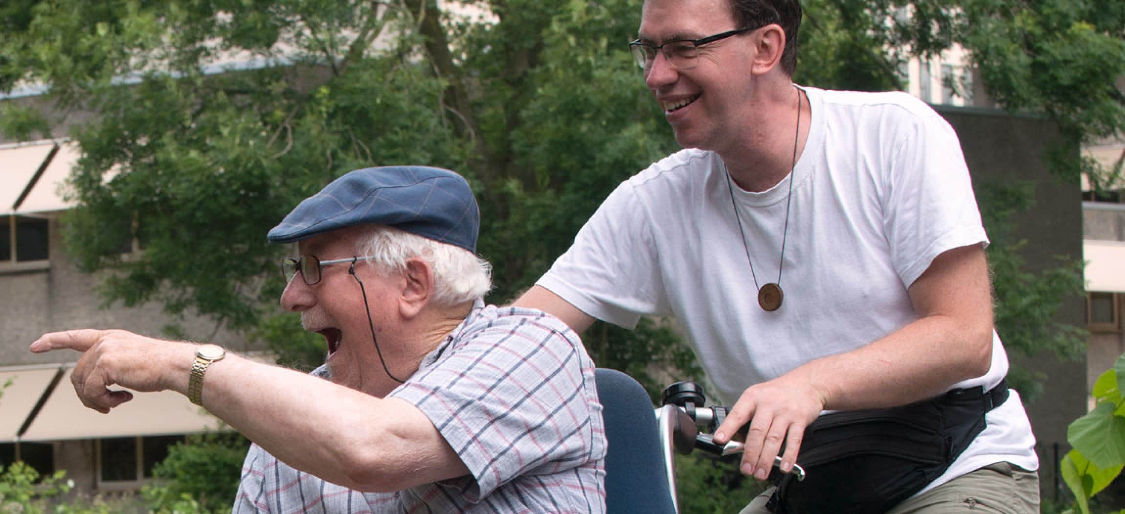 Enthousiaste blije man met klepmuts (ouderwetse pet) op een rolstoeltransportfiets met begeleider