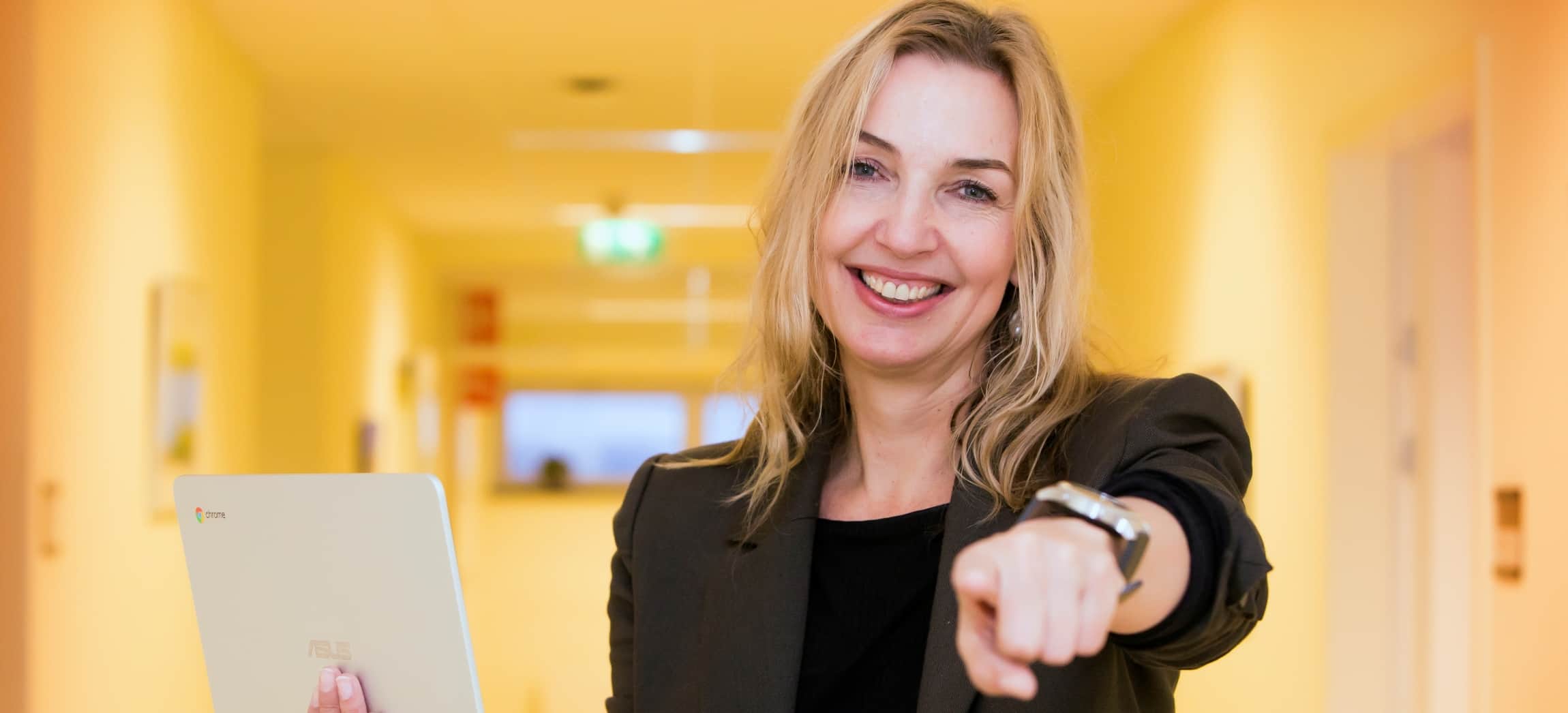 Vrouw met blonde haren in zwart colbertjasje houdt Chromebook vast en wijst naar de camera in gele gang
