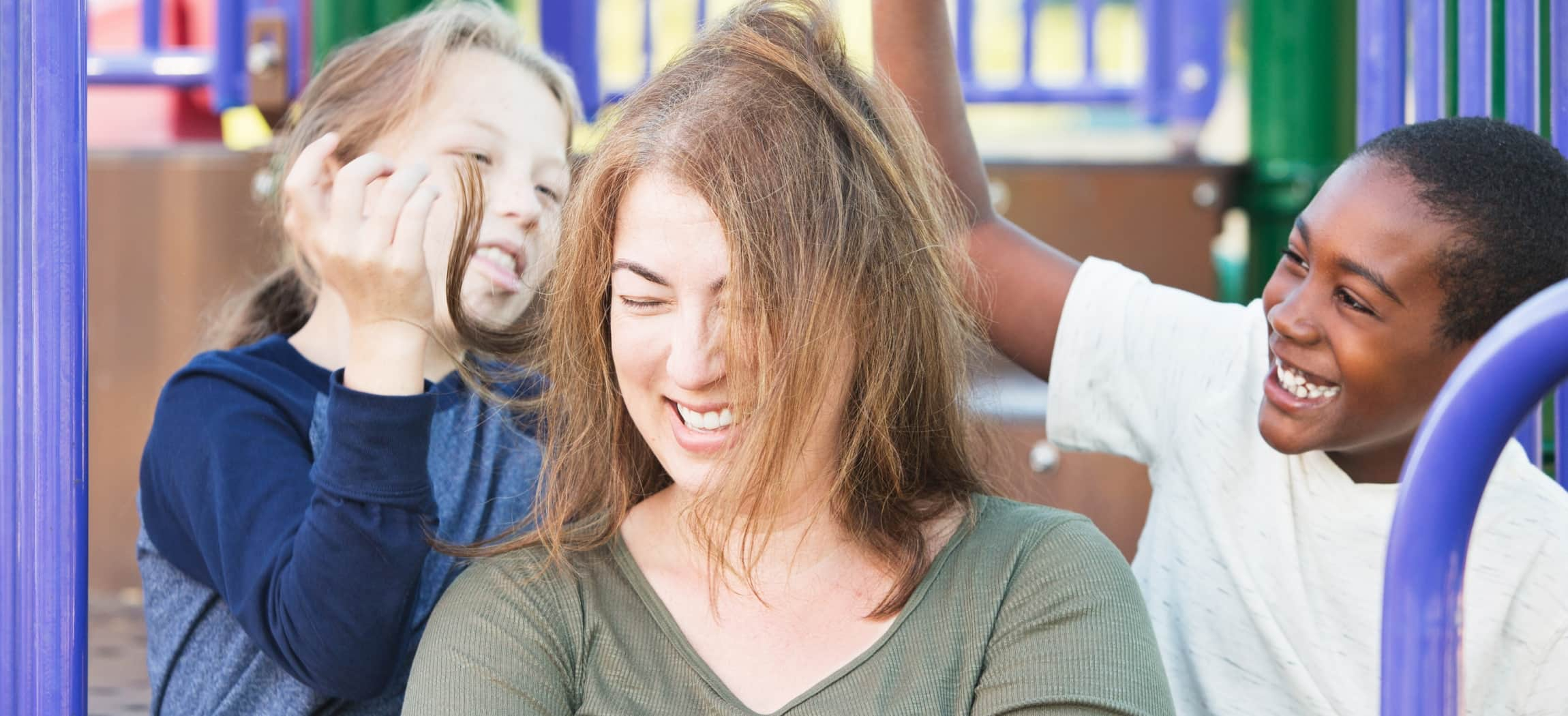 Lachende vrouw met warrig haar en 2 lachende kinderen op een buiten speeltoestel paars blauw