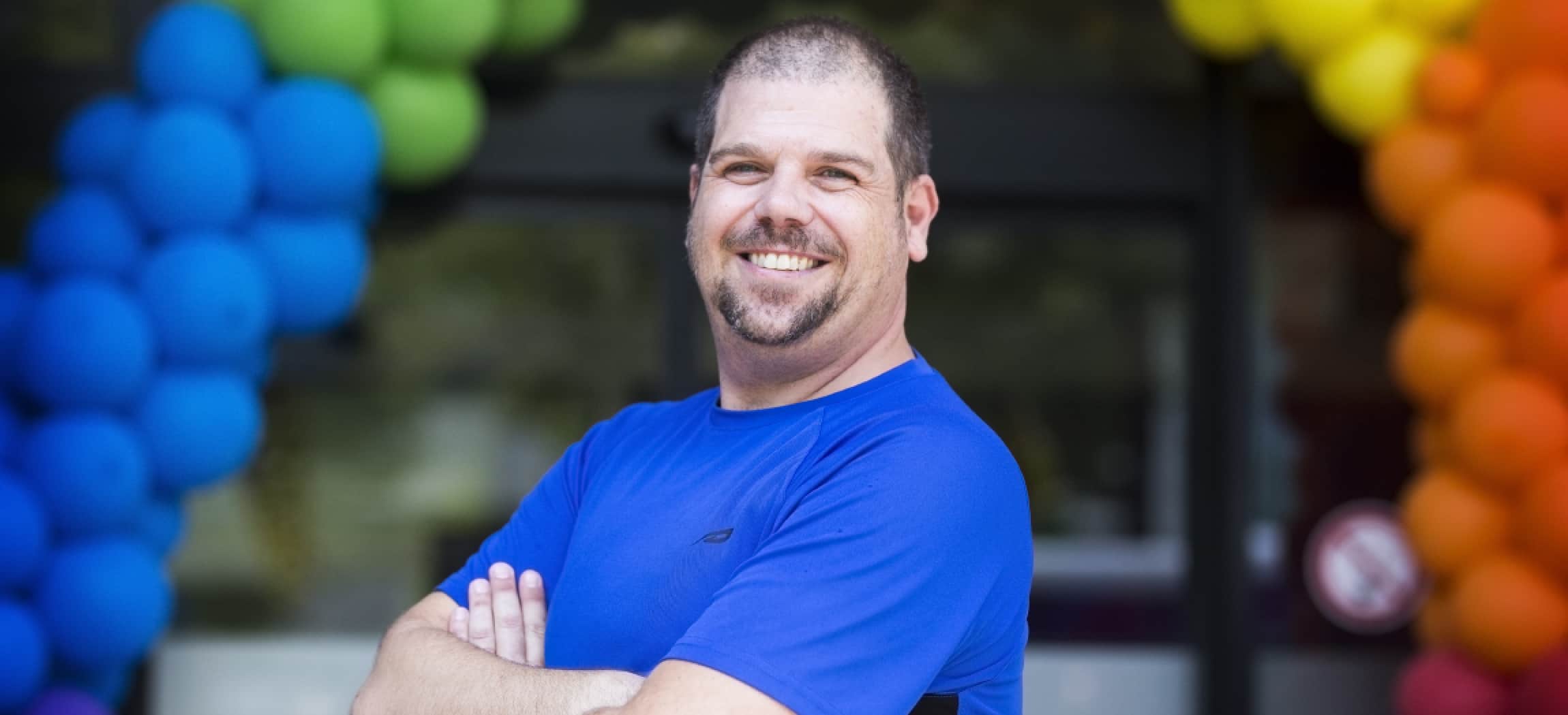 Jonge man met snor in blauw t-shirt, op achtergrond ballonnen-boog in regenboogkleuren Arjan Croonen