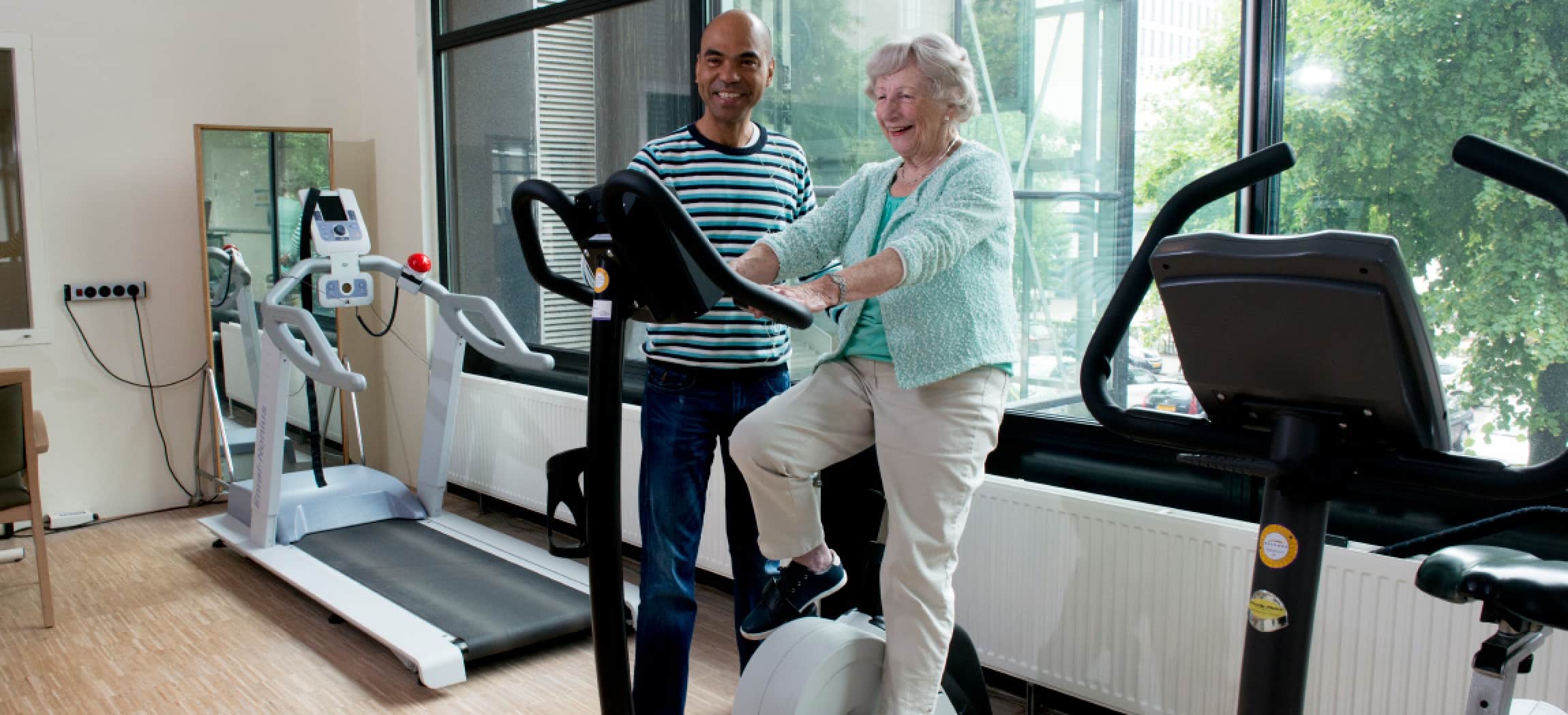 Ergotherapie oefenzaal fitness fiets oude vrouw met fysiotherapeut man