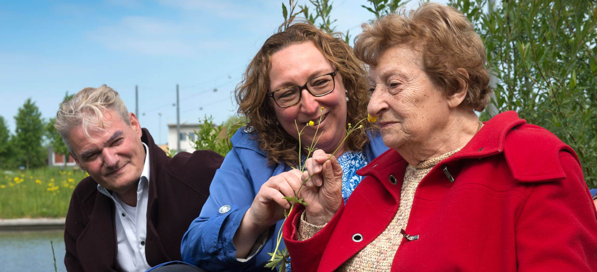 Zonnige dag buiten oudere vrouw met rode jas ruikt aan bloem en man kijkt toe