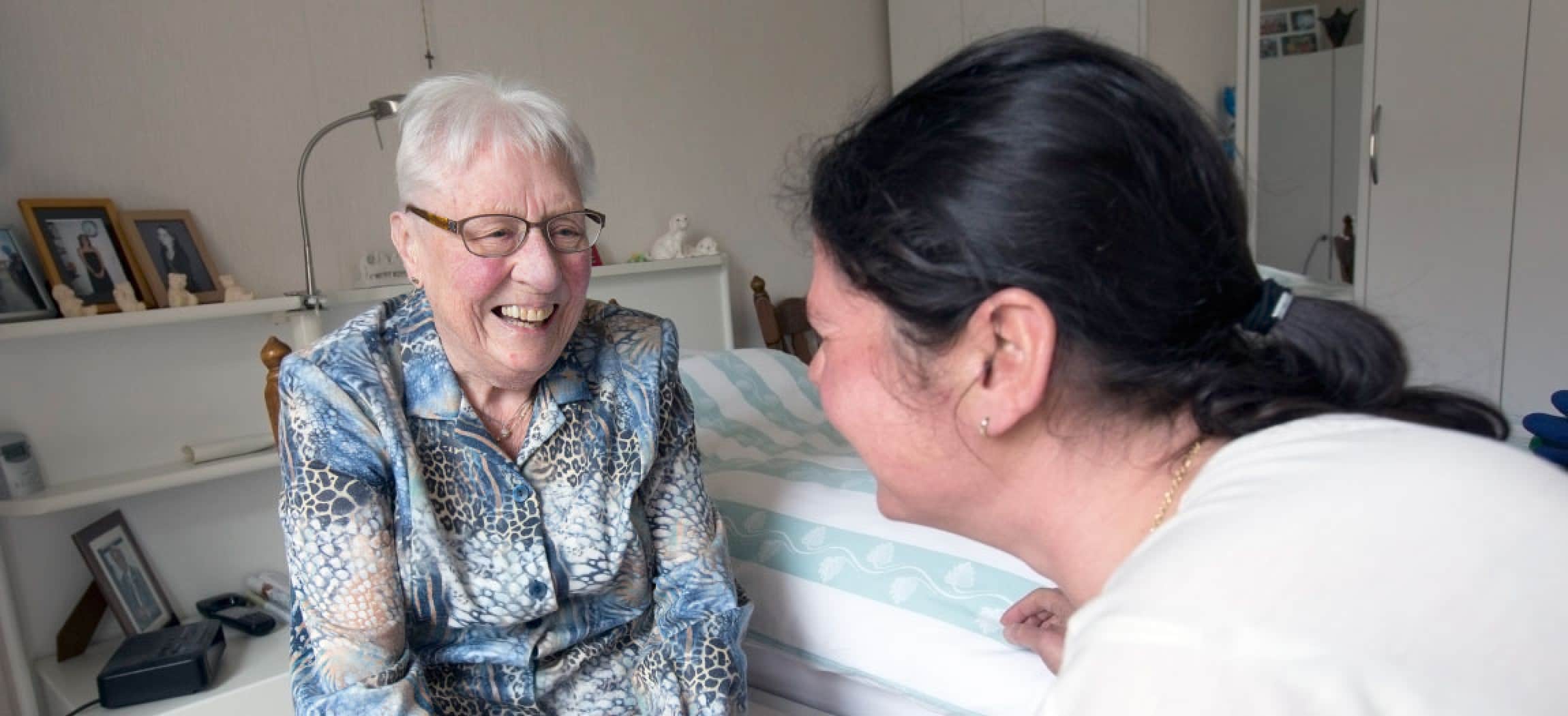 In slaapkamer van oudere vrouw kijkt wijkverpleegkundige oude vrouw aan