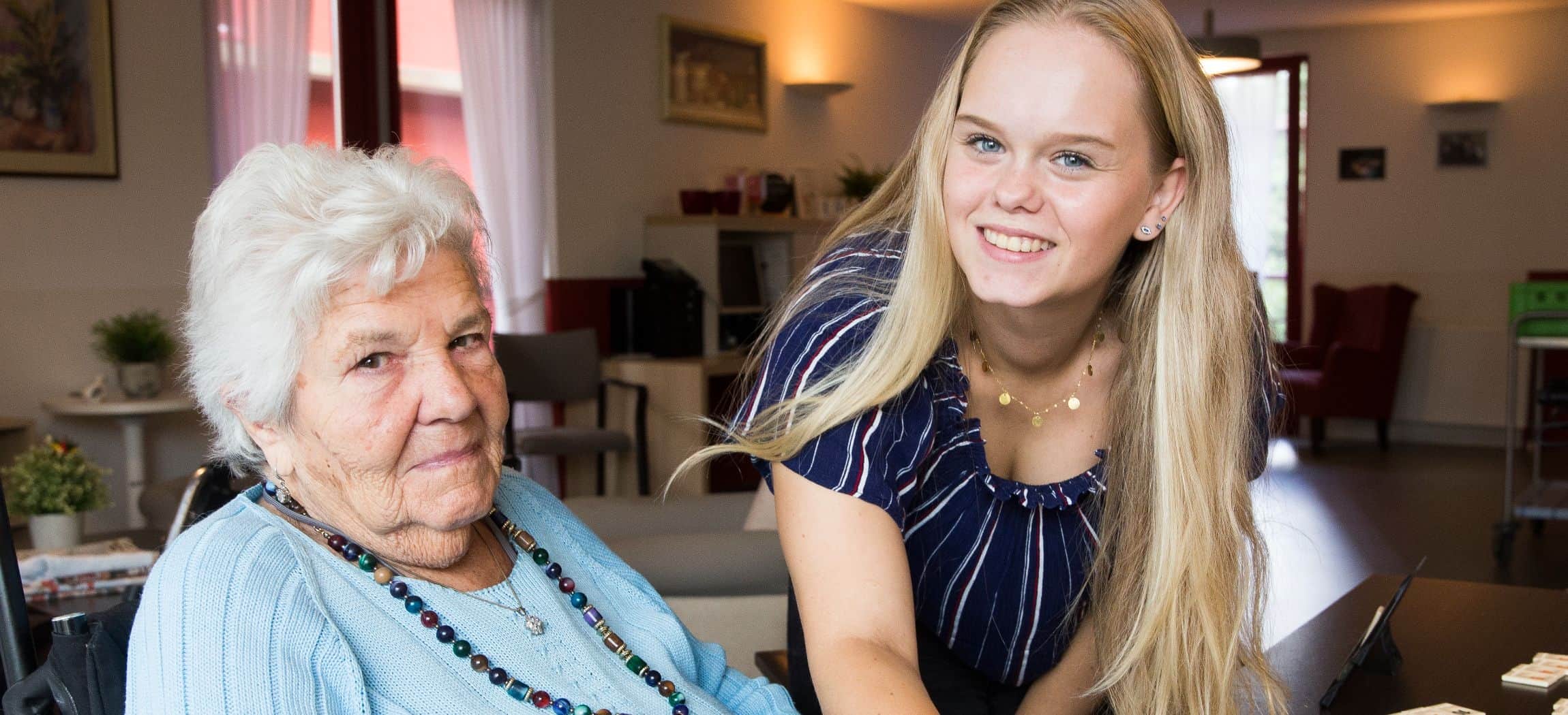 Jonge vrouw met lang blond haar, naast een bejaarde dame in lichtblauwe trui