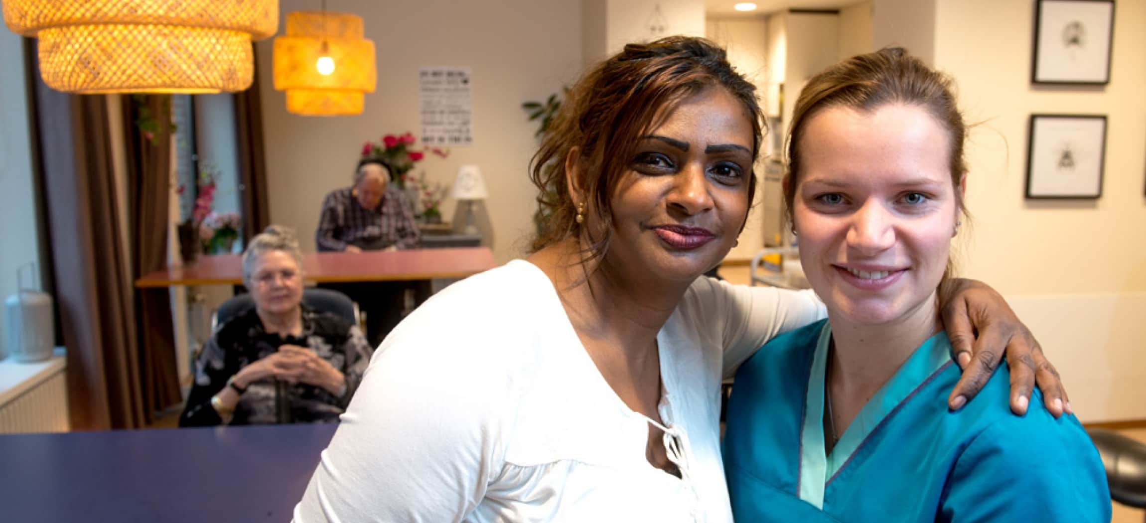 Zorgmedewerker in blauw uniform jasje wordt omarmt door collega in een bejaardenhuis