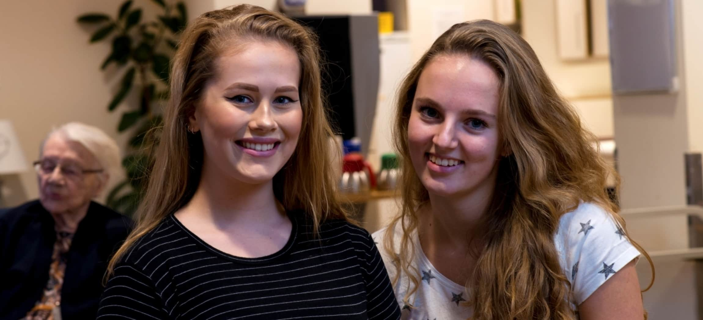 Twee jonge vrouwelijke stagiaires met blond haar, op de achtergrond een bejaarde vrouw