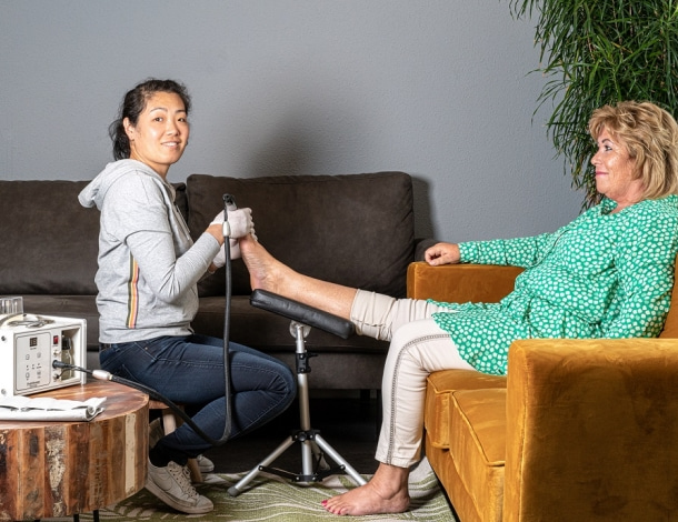 Vrouw met professioneel medisch pedicure toestel behandelt voet van vrouw zittend in fauteuil thuis