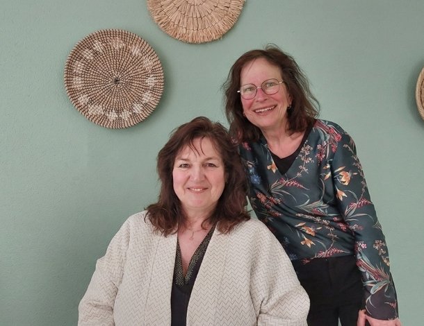 Twee vrouwen bekijken een boekje met poster Schijf van Vijf op achtergrond