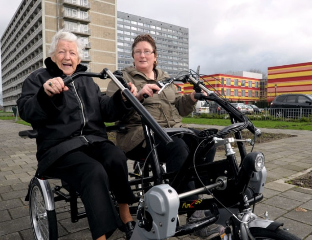 Duo-fiets buiten op de stoep met enthousiaste vrouw en op achtergrond gekleurd gebouw