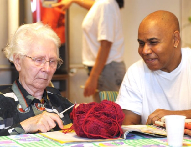 Mannelijke verzorgende kijkt naar oude vrouw met breiwerk en bol wol