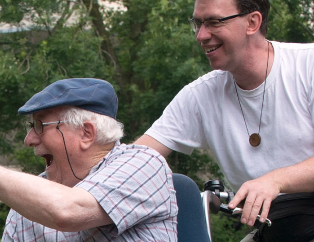 Enthousiaste blije man met klepmuts (ouderwetse pet) op een rolstoeltransportfiets met begeleider