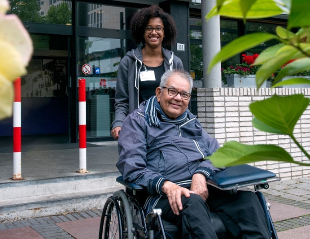 Man in rolstoel voor de deur van een verpleeghuis met een verpleegkundige
